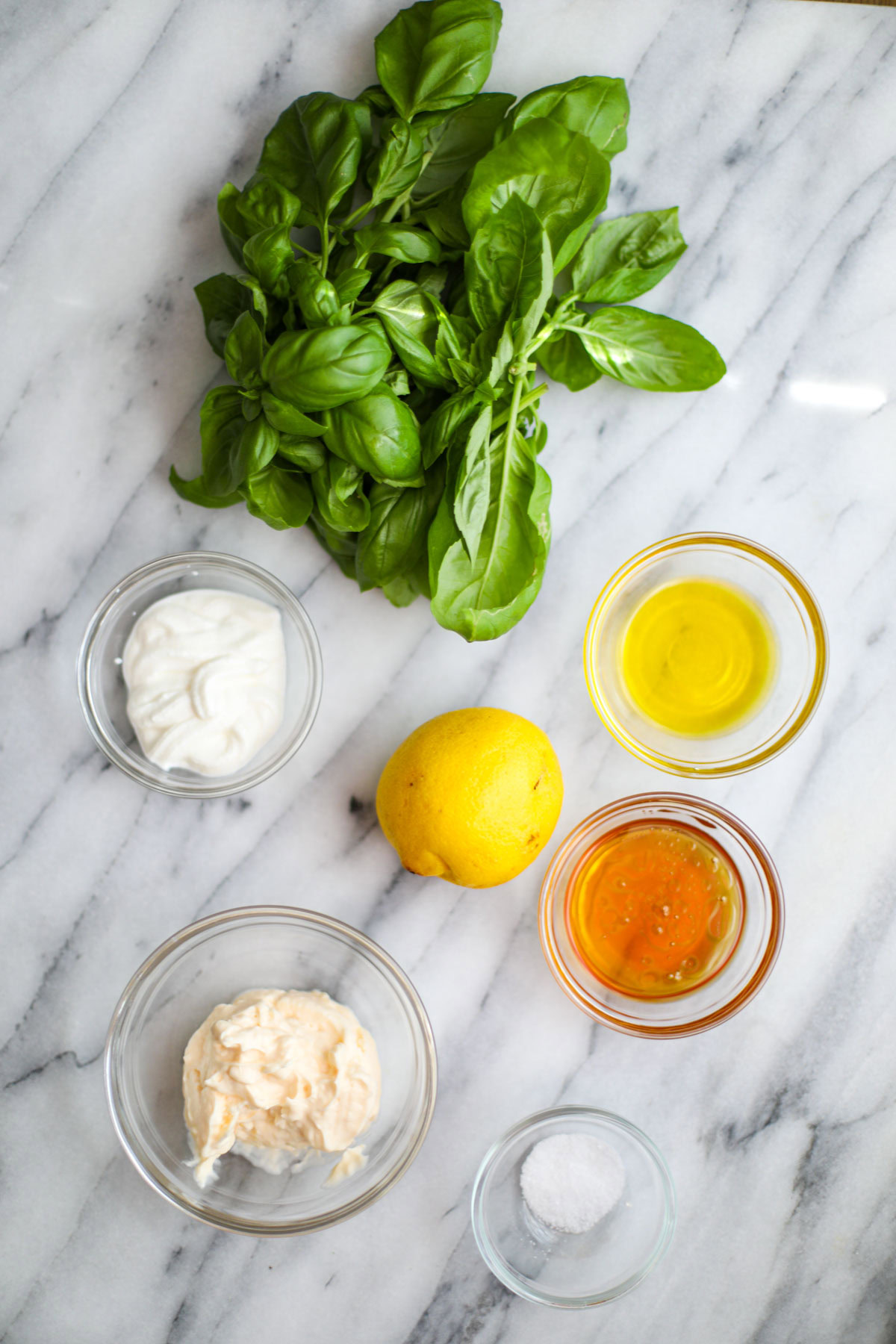 A bunch of basil, glass bowls of oil, honey, sour cream, mayonnaise, salt, and a lemon.