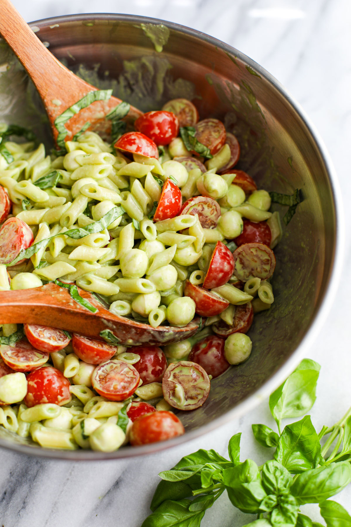 A stainless steel bowl of caprese pasta salad with wood salad servers. 
