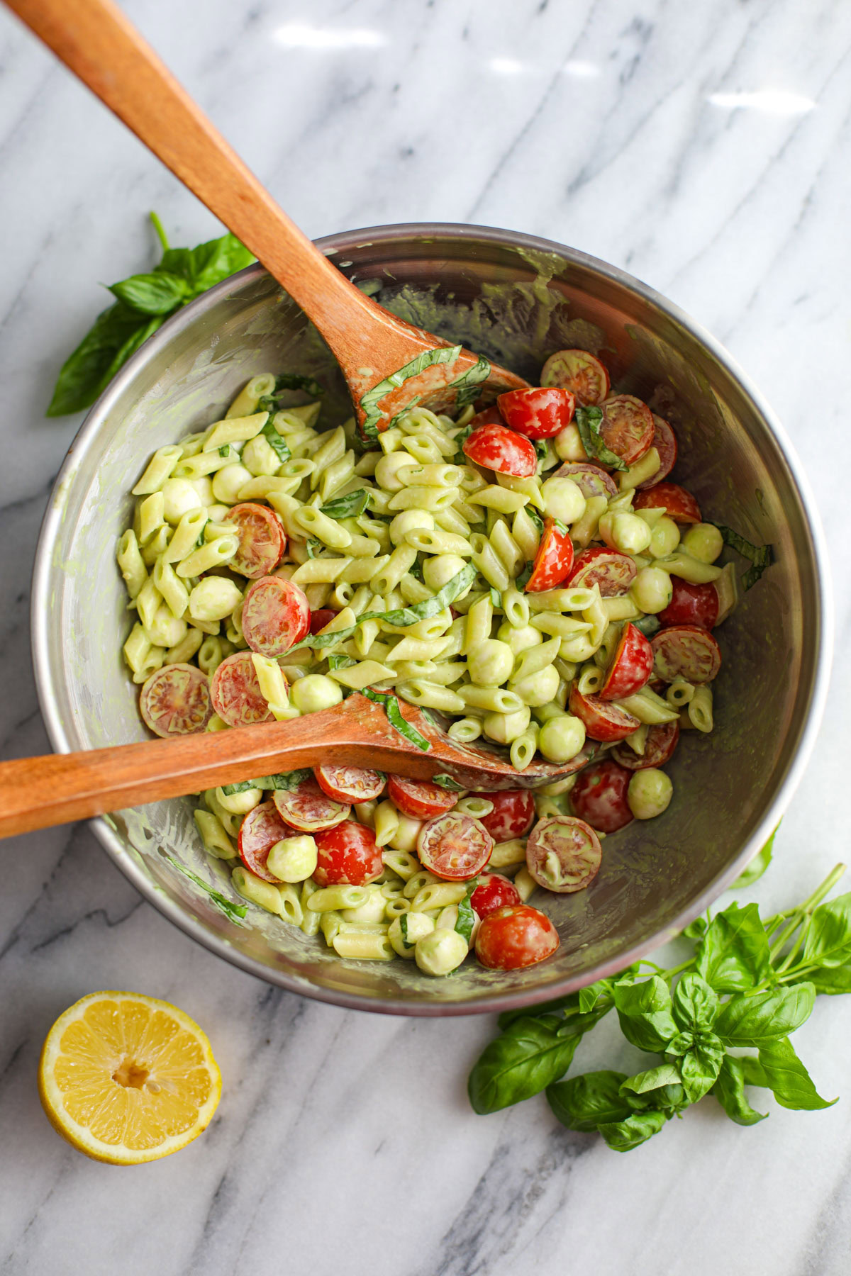 A stainless steel bowl of sweet basil caprese pasta salad with wood salad servers and basil on the side.