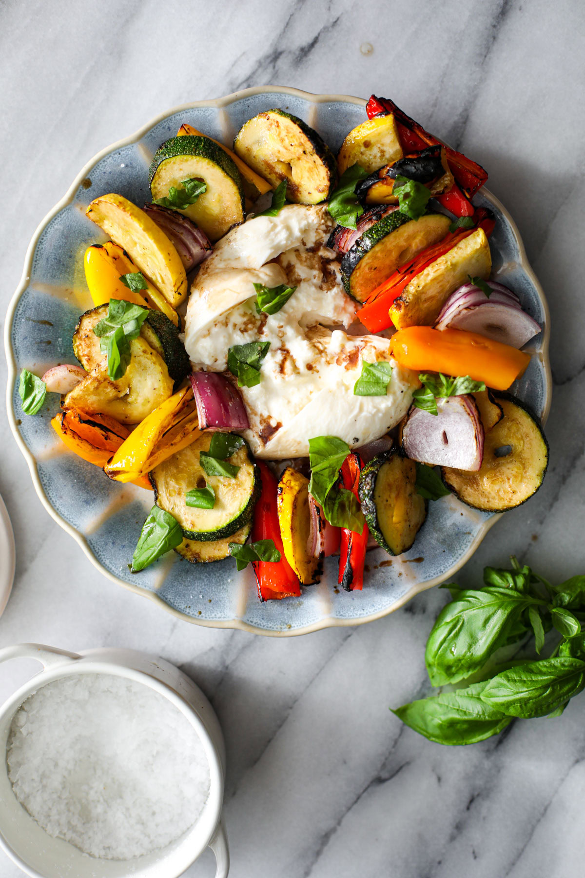 A blue scalloped plate with layered colorful balsamic marinated vegetables sitting around a broken ball of burrata with a bunch of basil next to it.