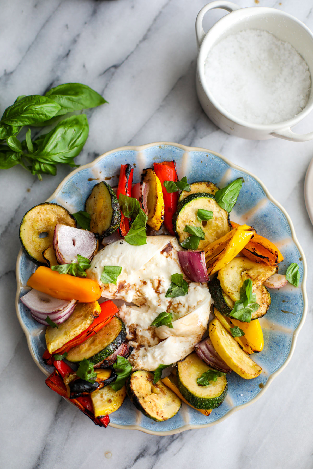 A blue scalloped plate with layered colorful vegetables sitting around a broken ball of burrata with a bunch of basil next to it.