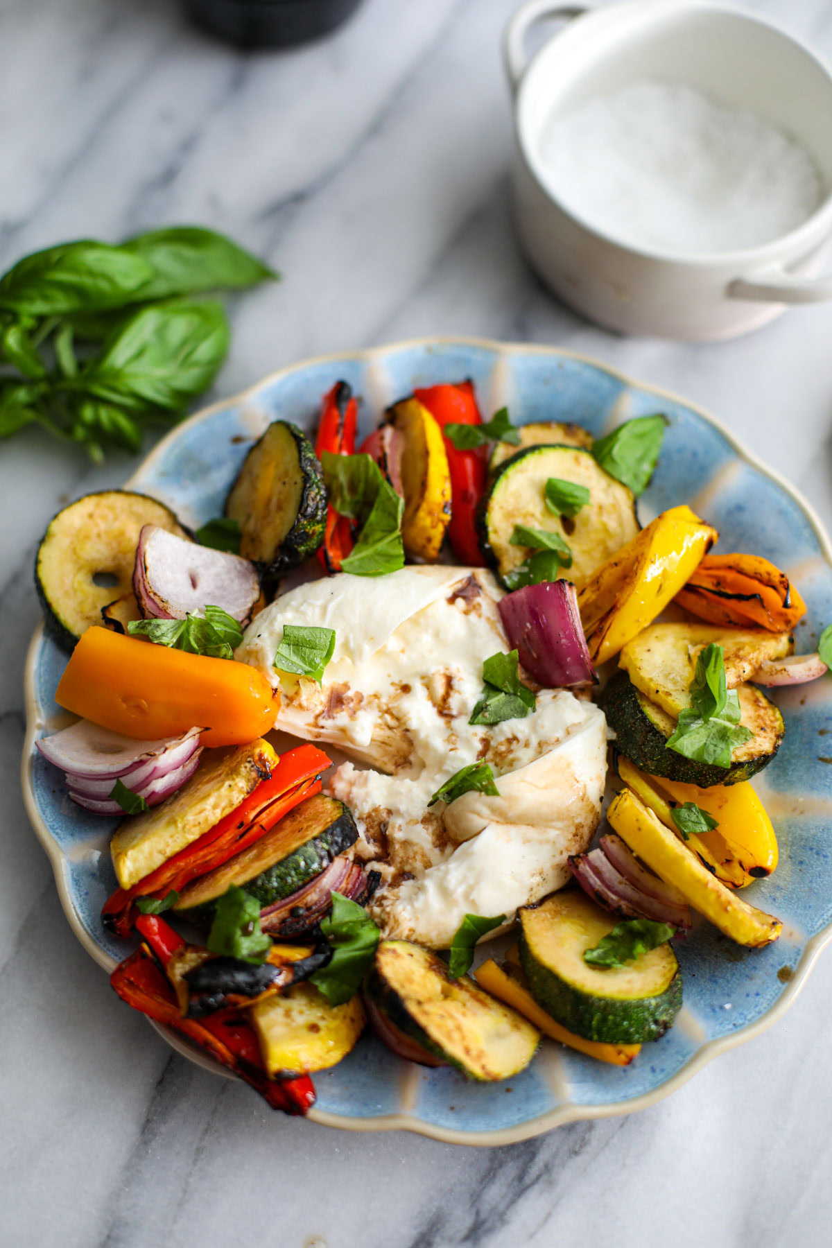 A blue scalloped plate with layered colorful balsamic marinated vegetables sitting around a broken ball of burrata with a bunch of basil next to it.