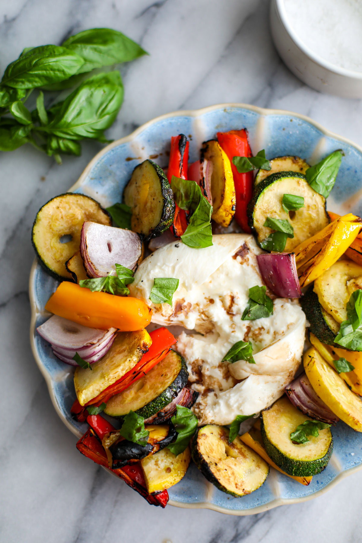 A blue scalloped plate with layered colorful vegetables sitting around a broken ball of burrata with a bunch of basil next to it.