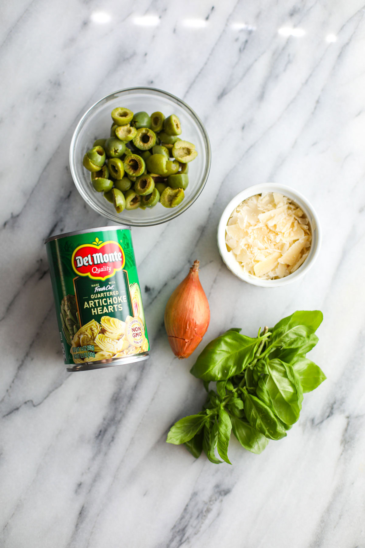 A glass bowl of olives, can of artichokes, bowl of cheese, bunch of basil and a whole shallot.