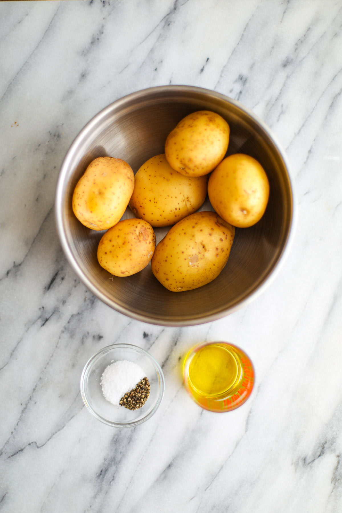 A bowl of potatoes, a container of olive oil, and a small bowl of salt and pepper.