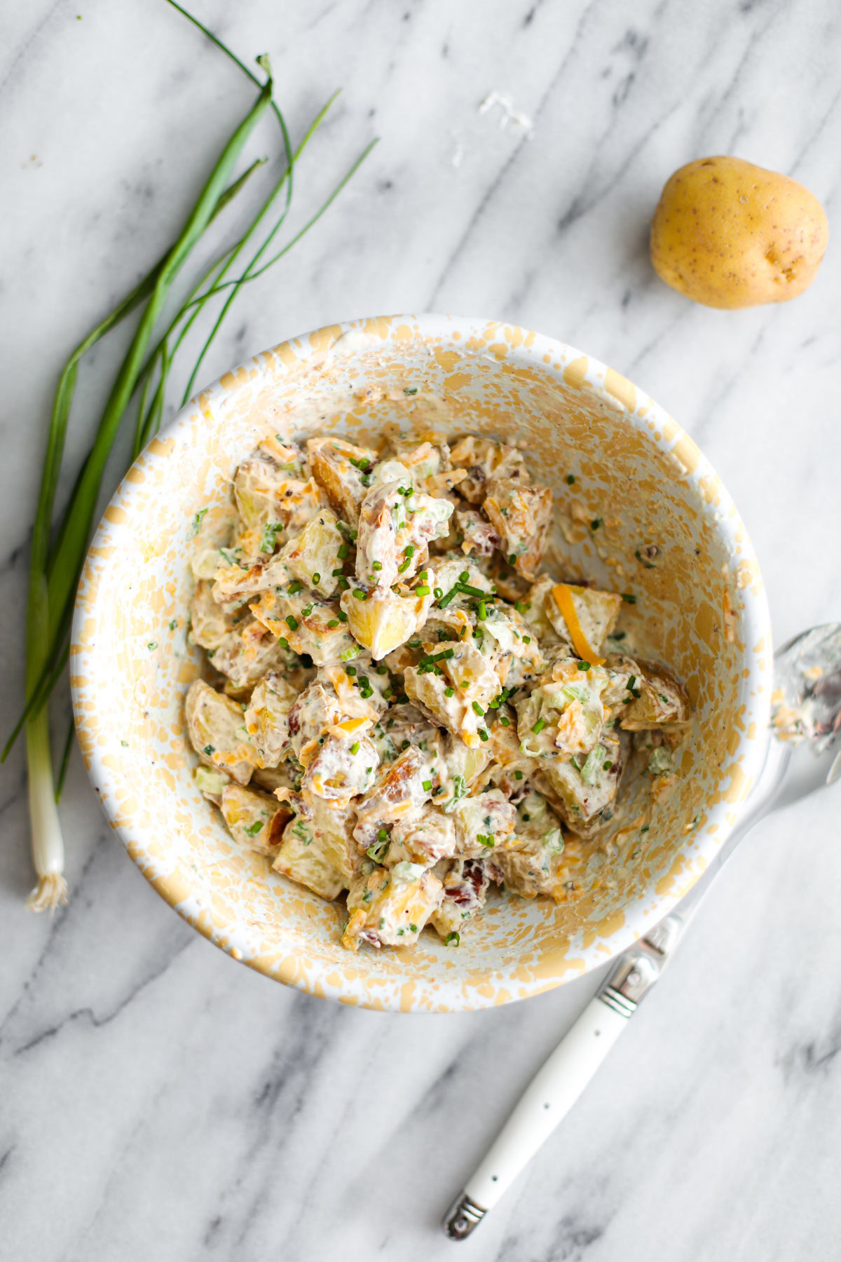 Baked potato salad in a white and yellow bowl with a potato, green onions, and a white spoon.