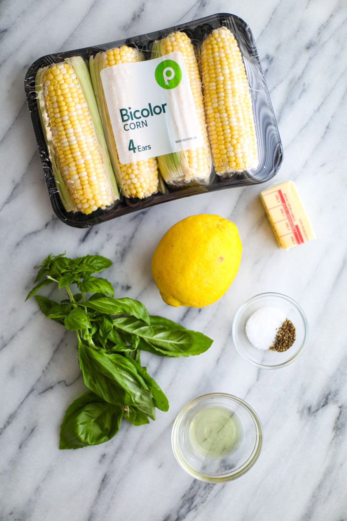 A package of corn, a half stick of butter, a lemon, a bunch of basil, a clear bowl of avocado oil, and seasonings.