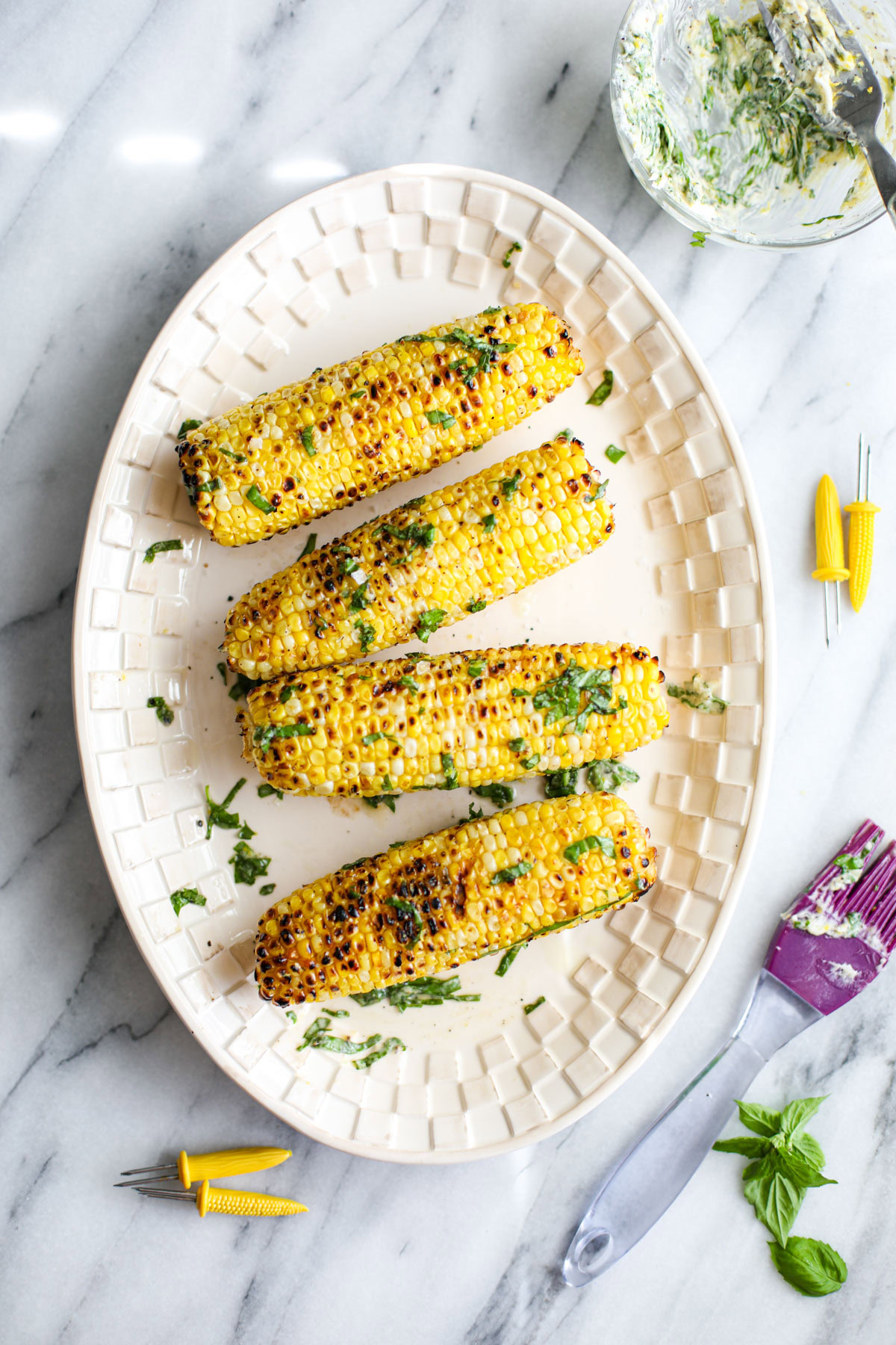 Basil Butter Grilled Corn on a white platter with a basting brush and bowl of compound butter.