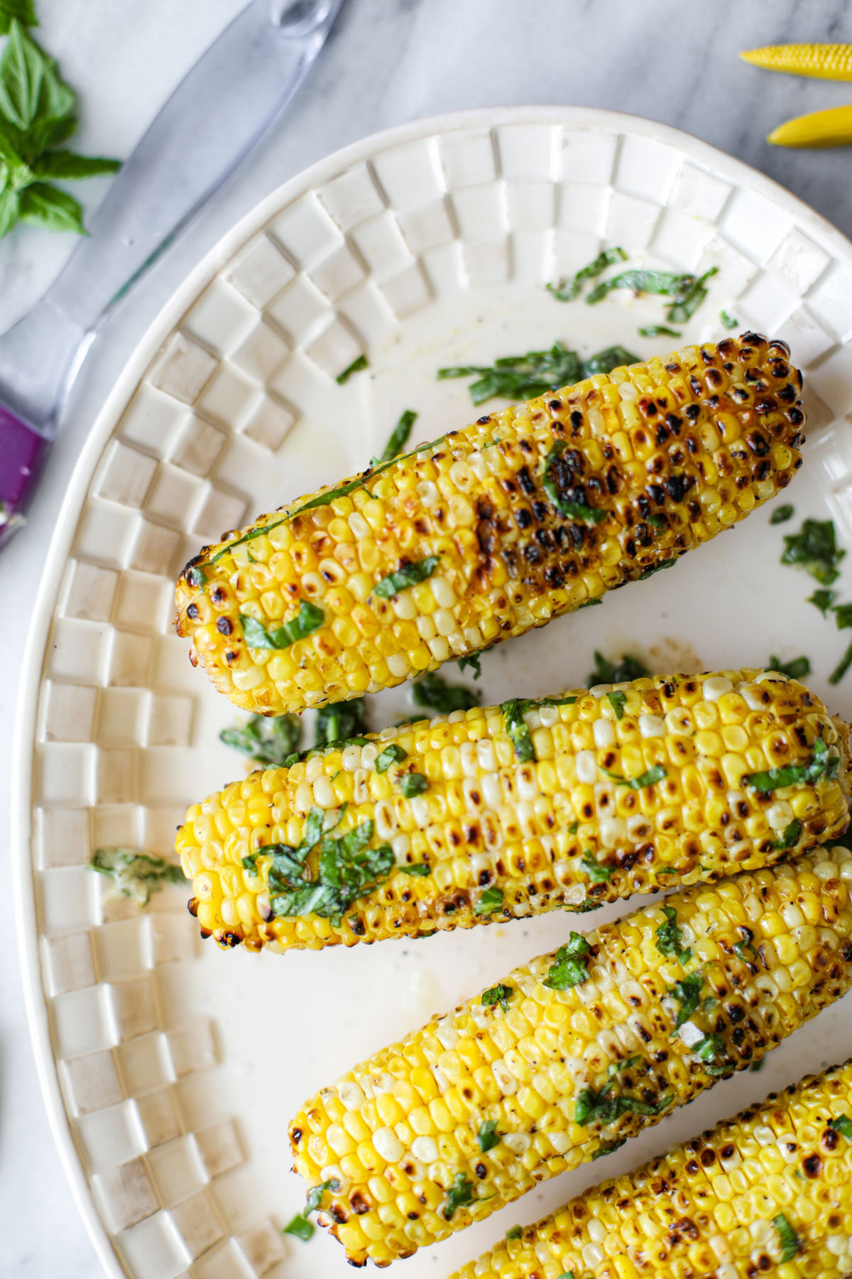 Basil Butter Grilled Corn on a white platter with a basting brush and bowl of compound butter.
