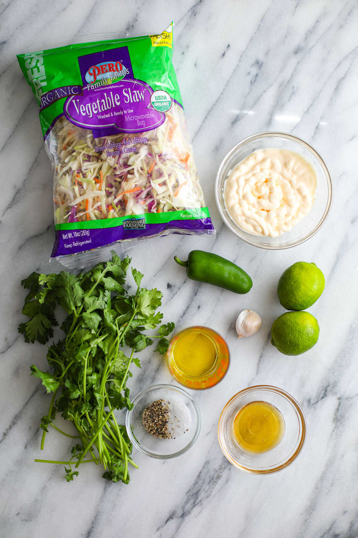 A bag of vegetable slaw, a bowl of mayonnaise, a jalapeño, a bunch of cilantro, two limes, a garlic clove, olive oil, honey, and seasonings all laid out on the counter.