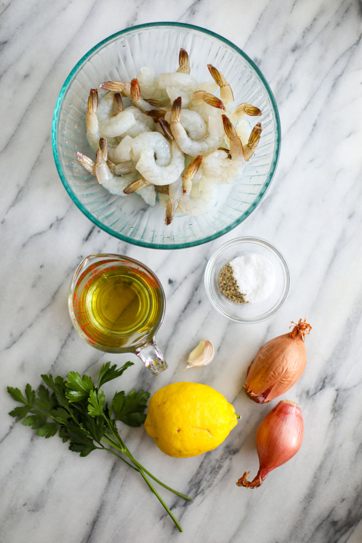 A bowl of raw shrimp, a measuring cup of oil, a glass bowl of seasonings, 2 shallots, a lemon, a garlic clove, and a bunch of parsley.