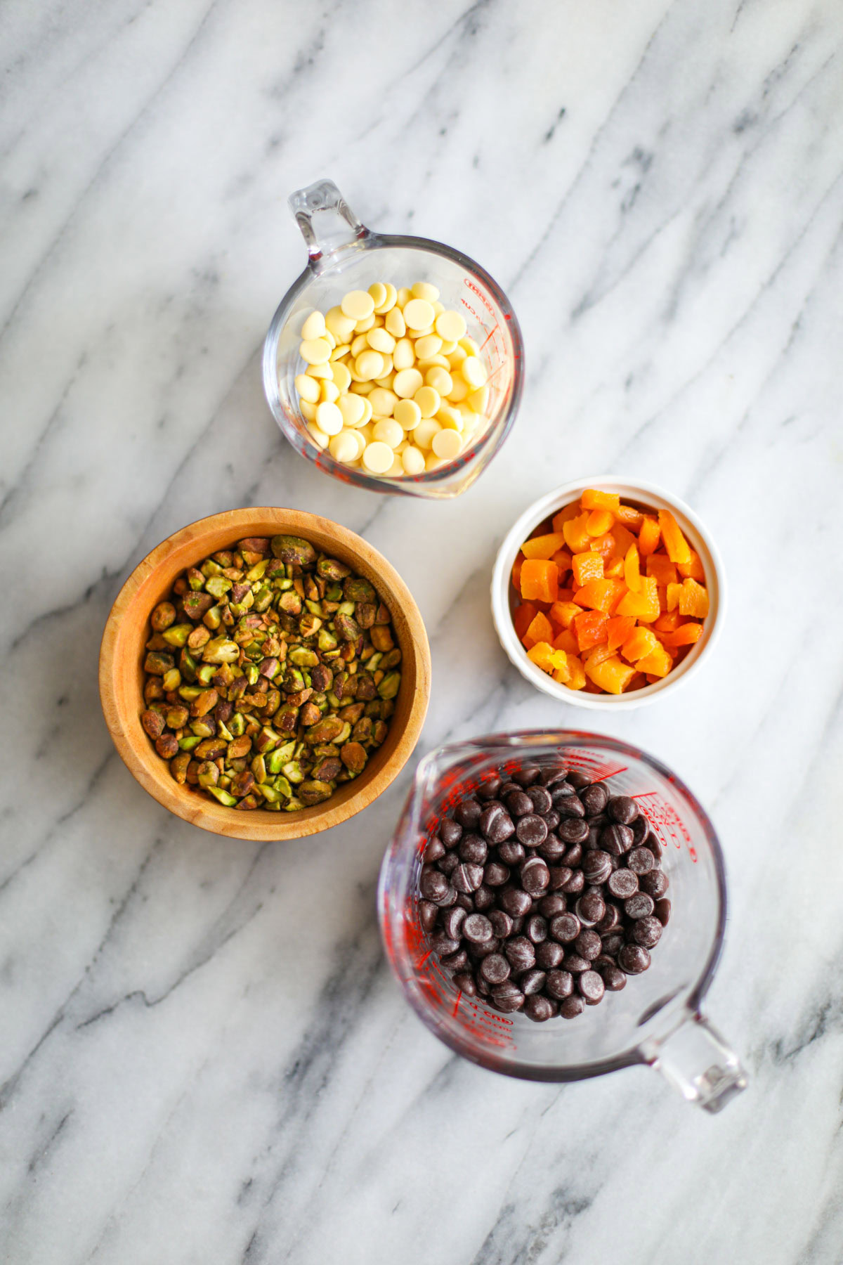 Bowls of chocolate chips, chopped pistachios, and chopped apricots.
