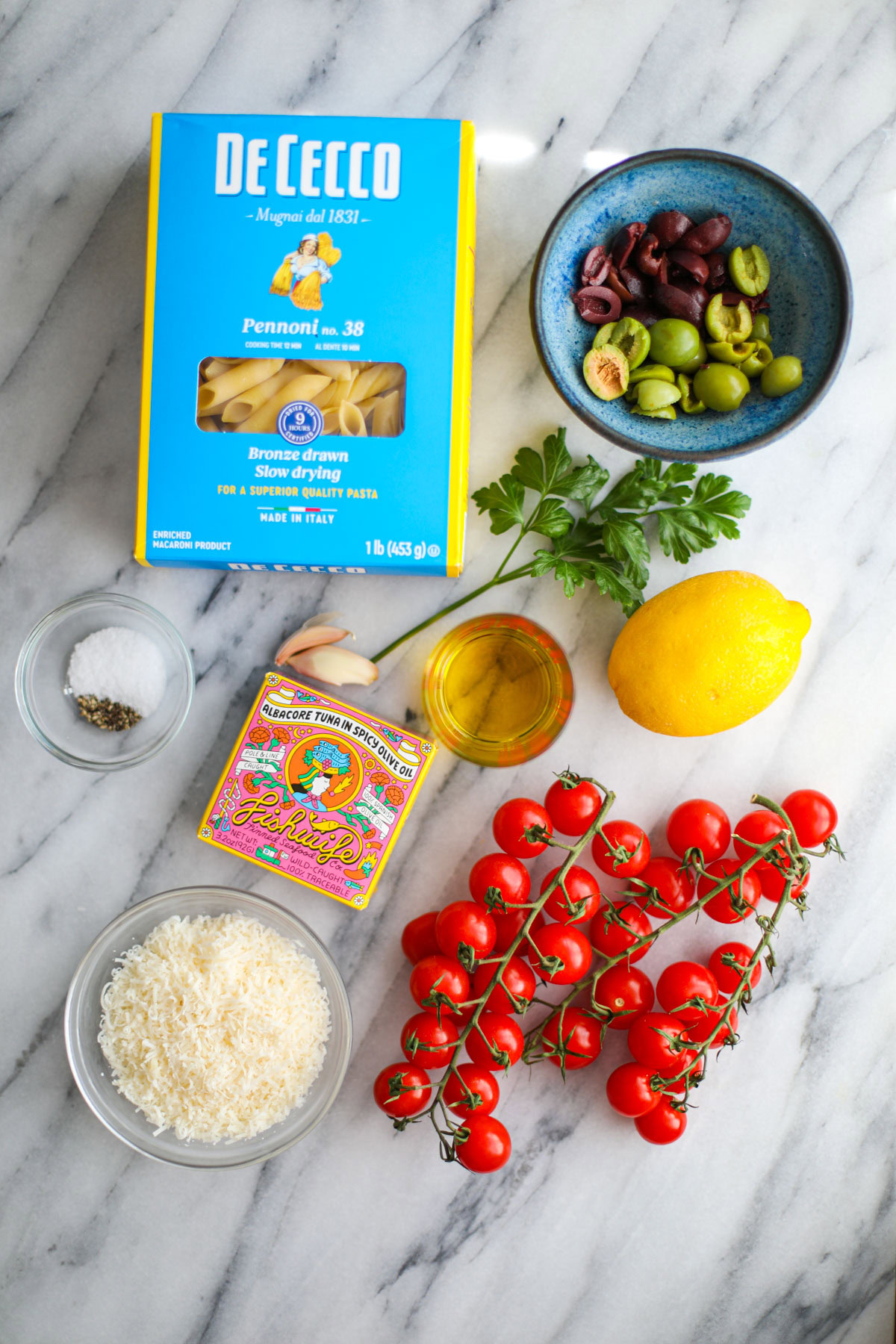 A blue box of pasta, bowls of olives, oil, parmigiano, 2 cherry tomatoes on the vine, a lemon, a can of tuna, two garlic cloves, and a bunch of cilantro.