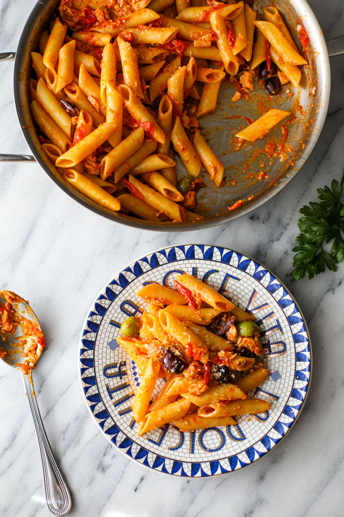 Canned tuna pasta in a stainless skillet with a blue and white plate served with extra olives and parmesan.
