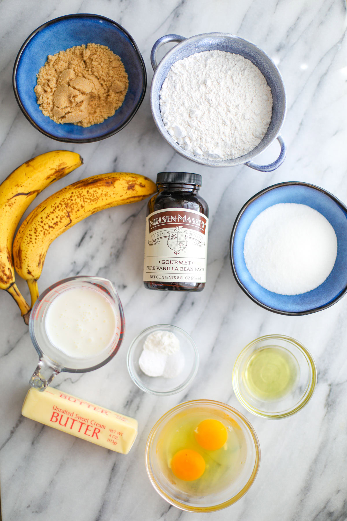 Ingredients to make banana cake including cake flour, bananas, vanilla bean paste, buttermilk, white sugar, oil, baking soda, eggs and butter.