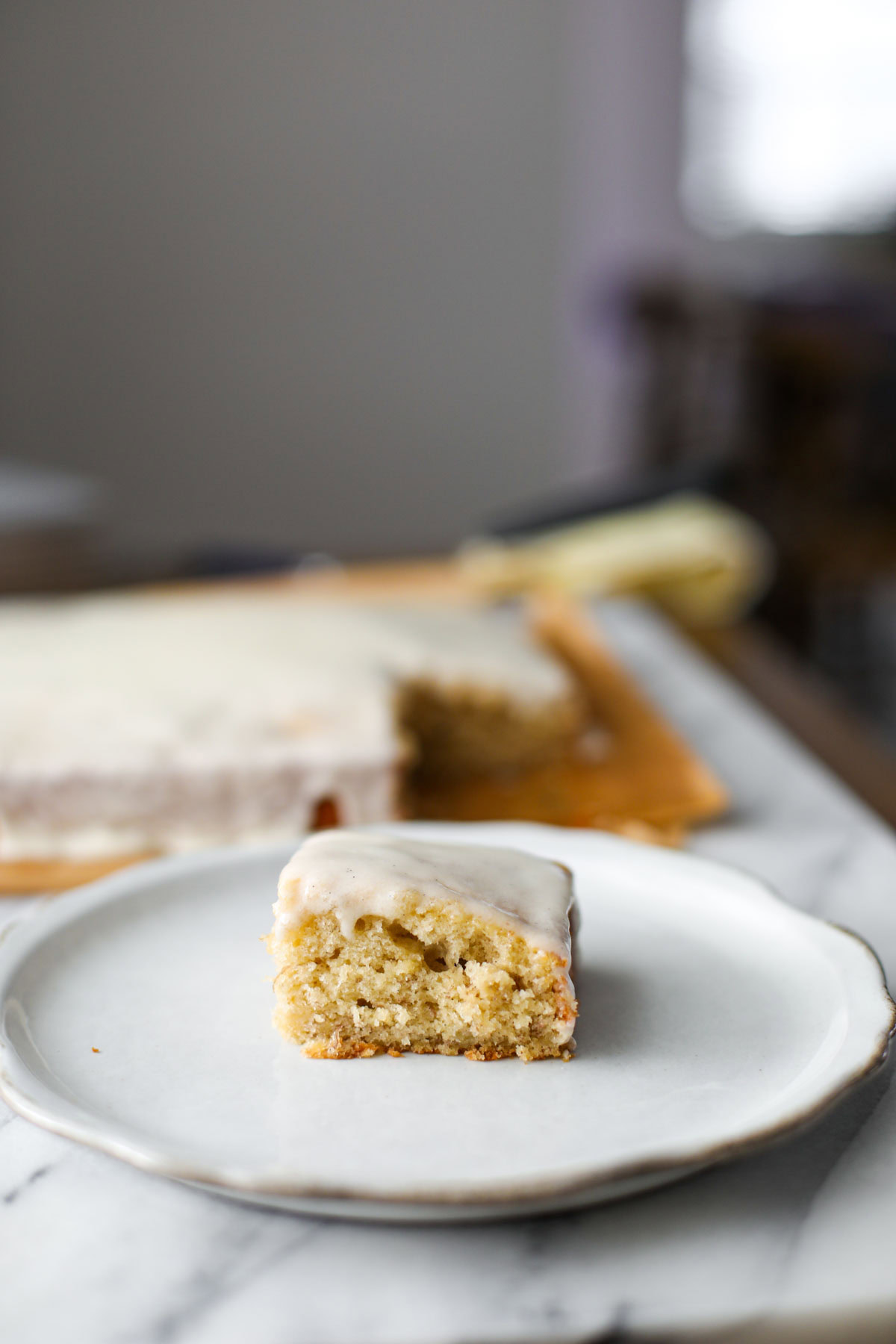 A slice of banana cake with vanilla bean icing on a white plate.