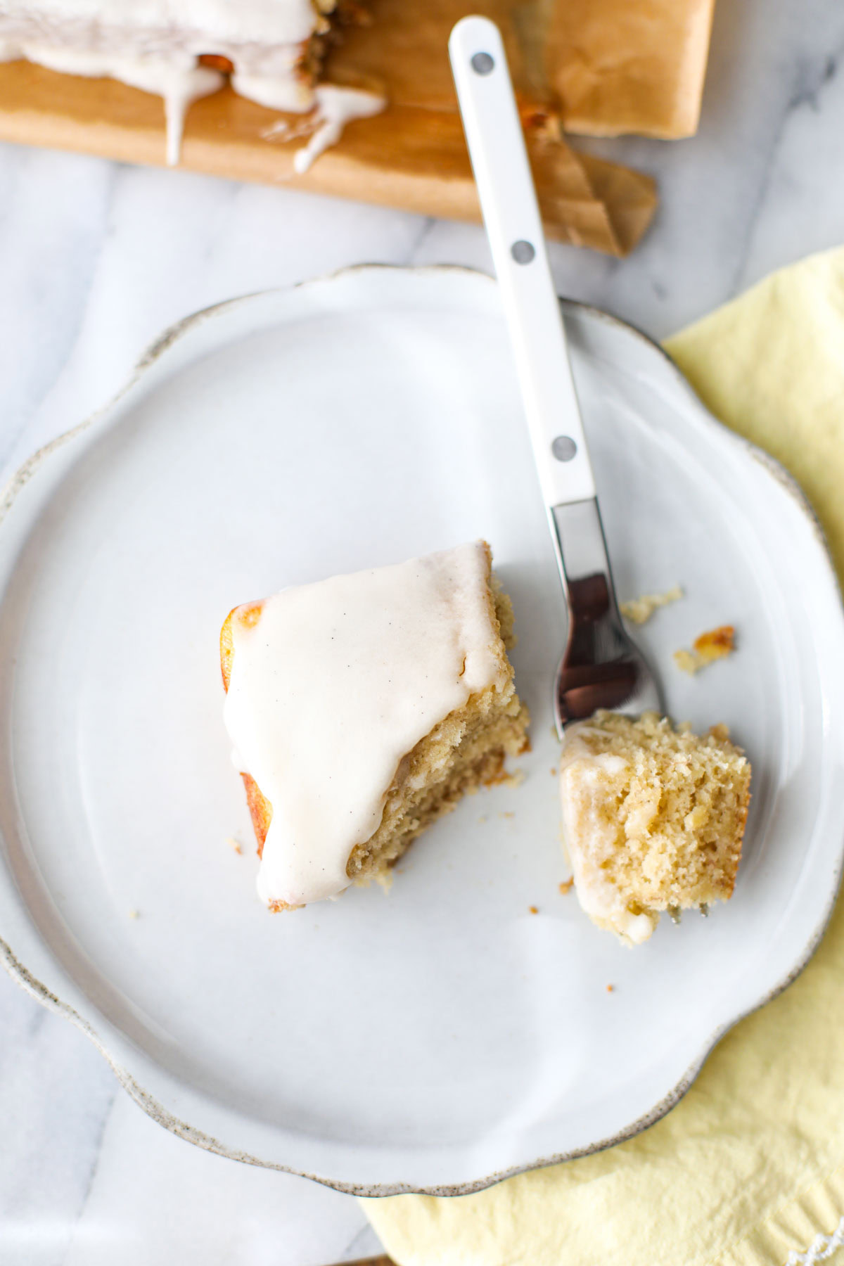 A slice of banana cake with vanilla bean glaze on a white plate with a white fork.