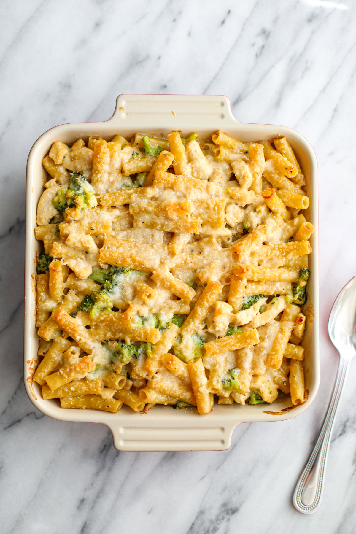 A square baking dish of chicken broccoli ziti with a silver serving spoon.