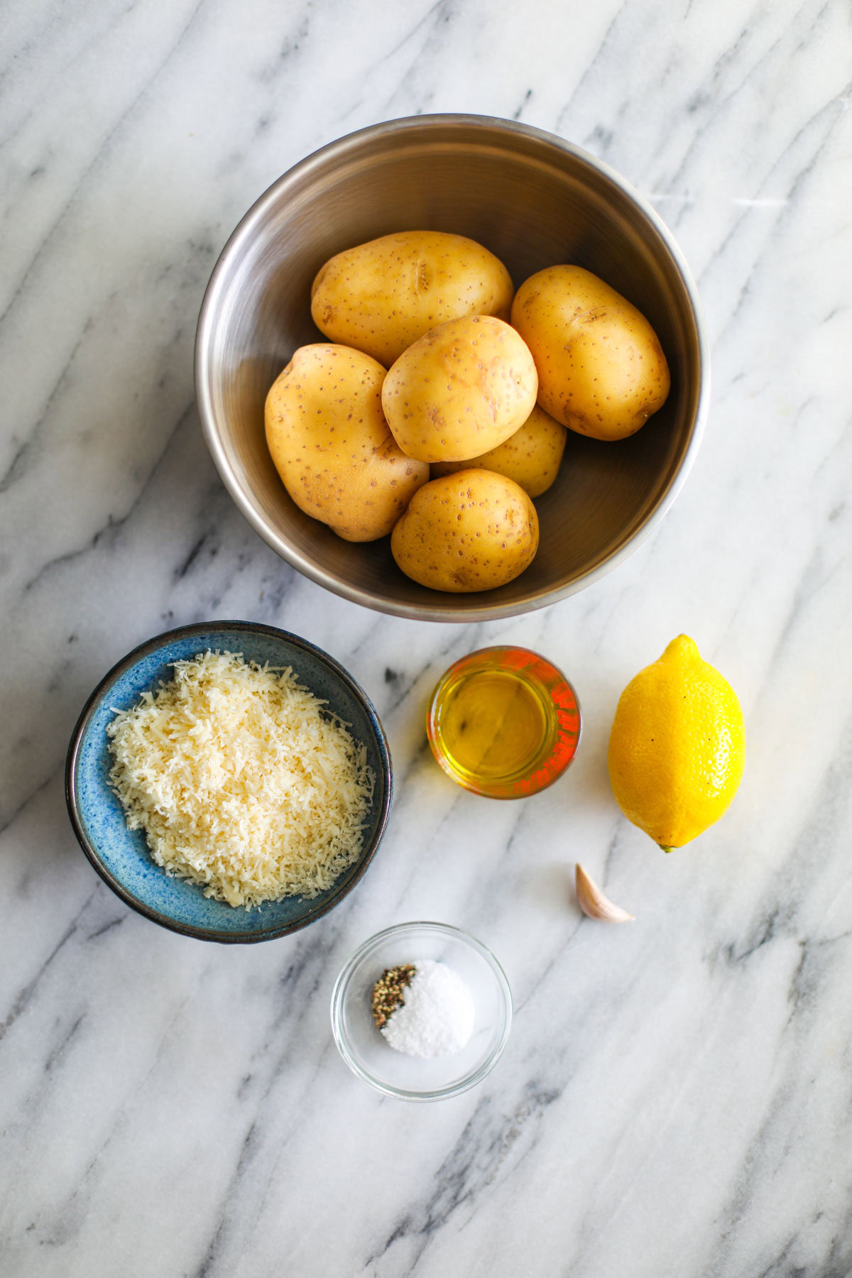 A stainless steel bowl of gold potatoes, a bowl of parmigiano reggiano, a whole lemon, a measuring cup of oil, a garlic clove, and a glass bowl of seasonings.