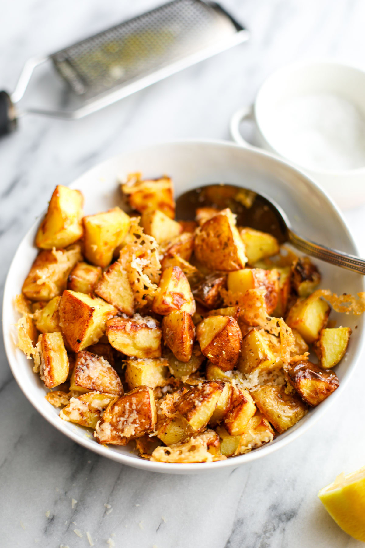 A white bowl of crispy parmesan potatoes on a marble counter with a silver spoon.