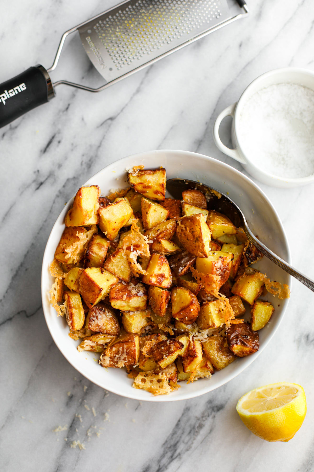 A white bowl of crispy parmesan potatoes on a marble counter with a silver spoon and a microplane zesting tool.