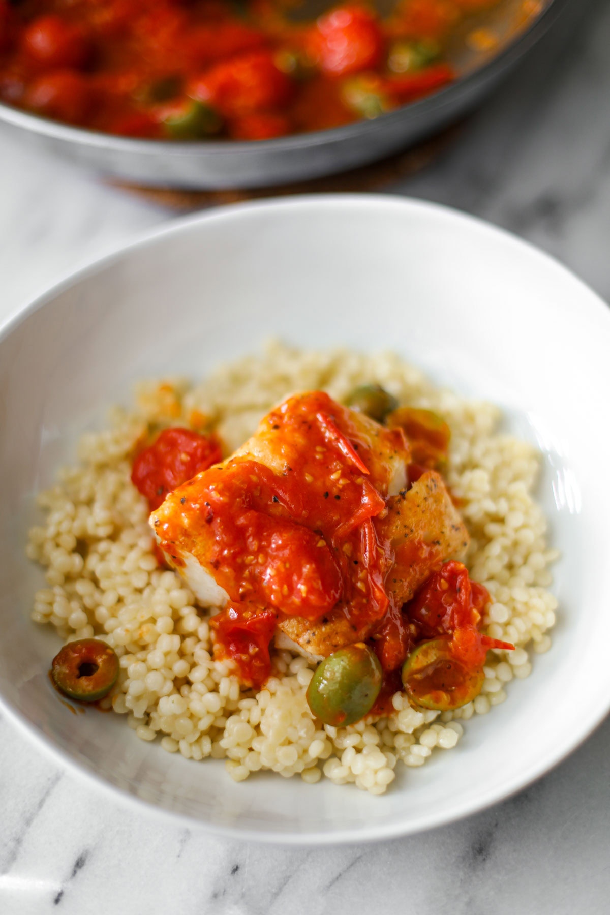 A white bowl of couscous with pan seared cod and jammy cherry tomatoes.
