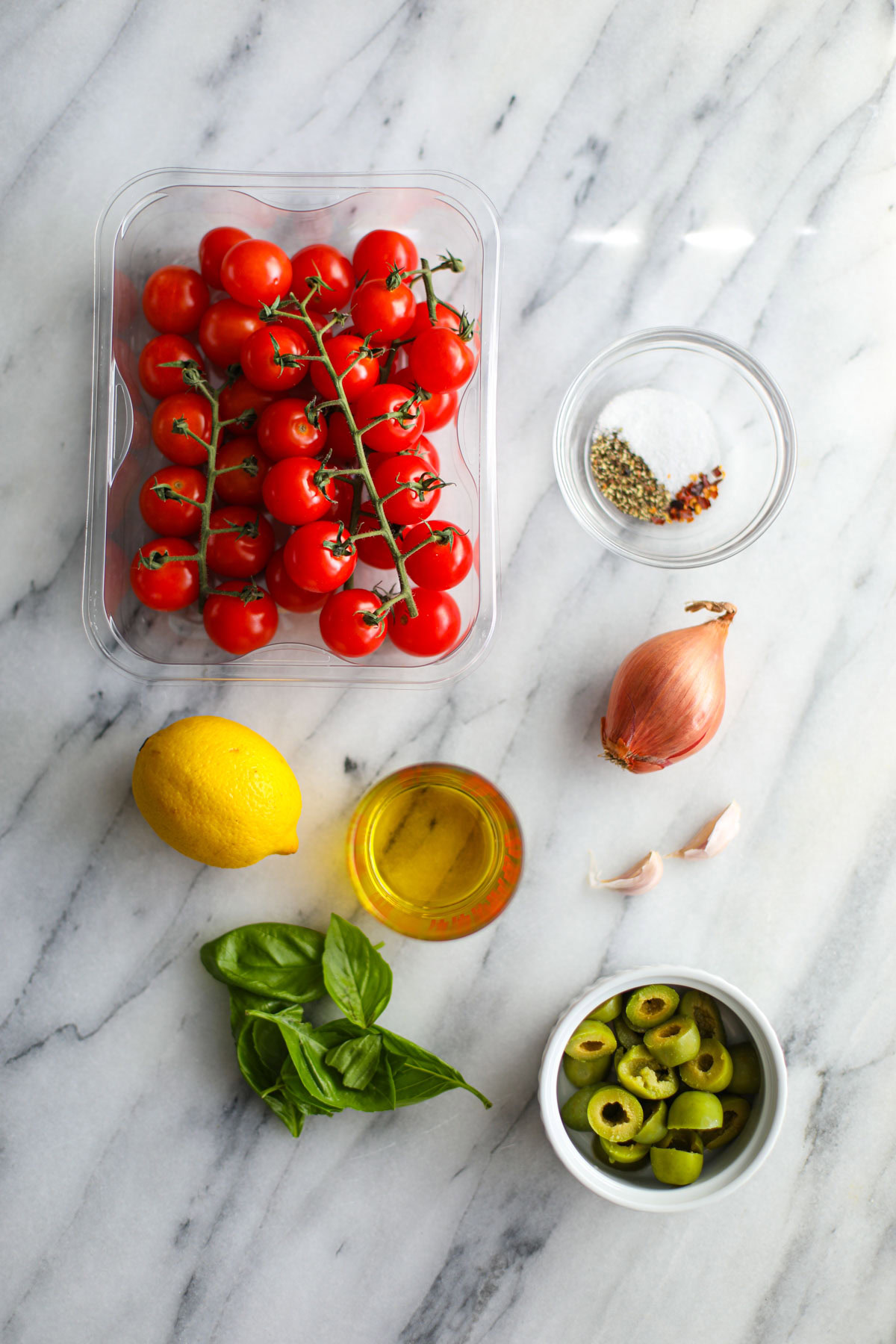 A container of cherry tomatoes, seasonings, a shallot, lemon, basil, garlic, and olive oil.