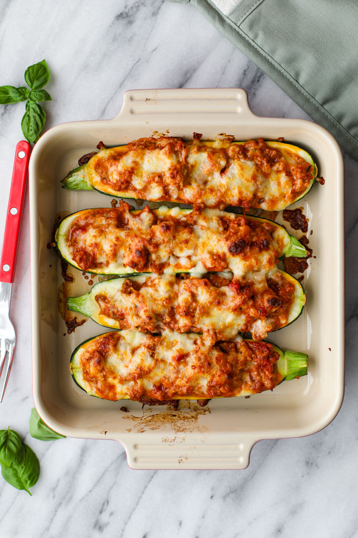 Sausage stuffed zucchini boats in a square baking dish with a red fork and pieces of basil.